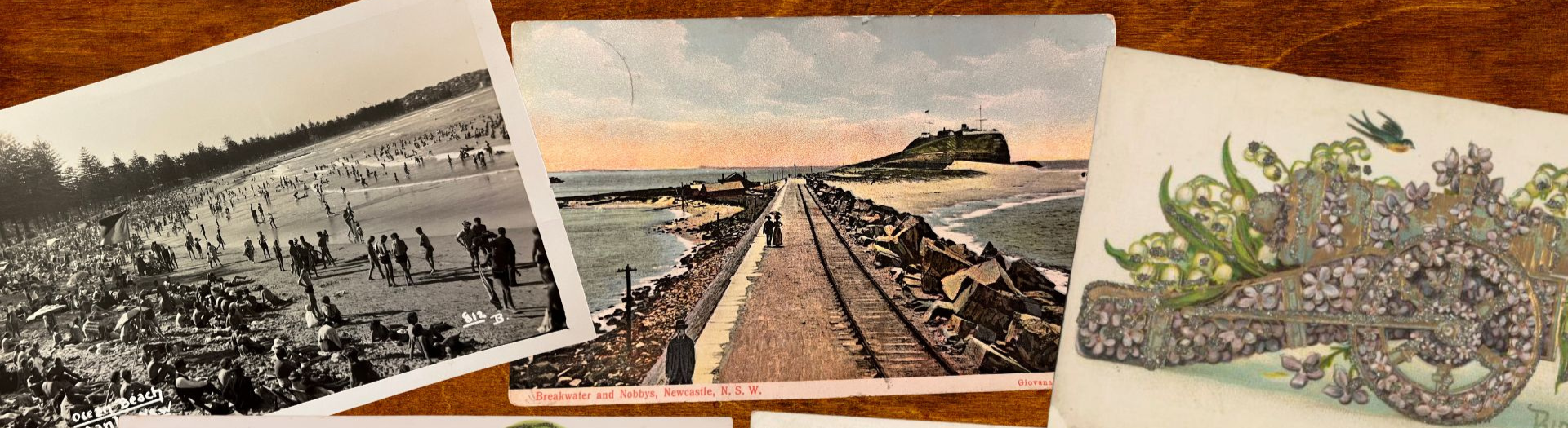 Three old postcards on a wooden surface. From right to left, they show a crowded beach, a couple walking along a pier and a cannon covered in flowers