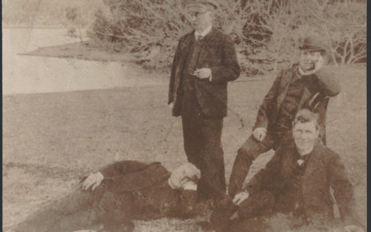 A black and white photograph of three men in suits and waistcoats. One man is standing another is sitting on a chair, another is sitting on the grass and the fourth is laying at the feet of the standing man. They are outside and positioned in front a some large bushes.
