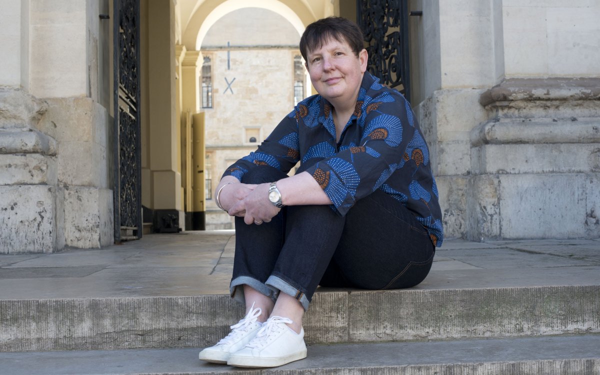 A woman sitting at the top of some stairs with her arms wrapped around her legs. She has short hair and is wearing a blue top, dark pants and white sneakers.