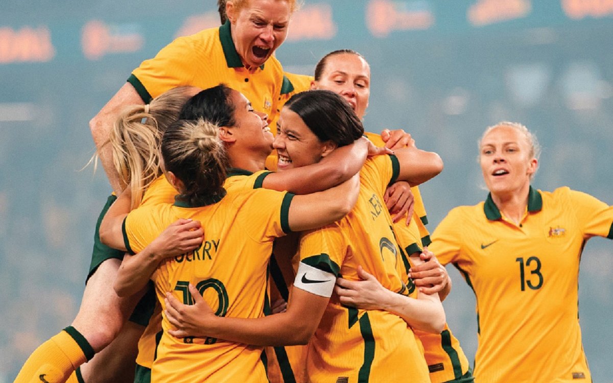 Seven women in the Australian soccer uniform all cheering and hugging one another.