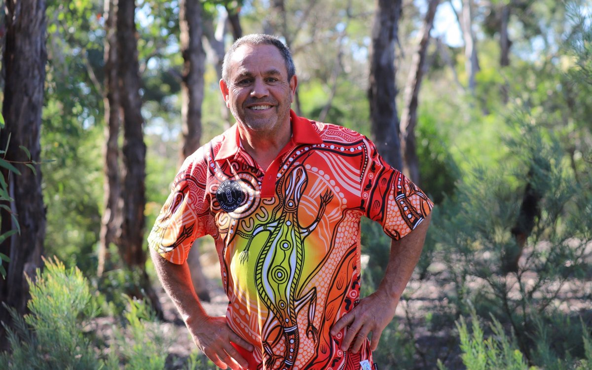 A man wearing a colourful shirt, standing amongst trees and shrubs.