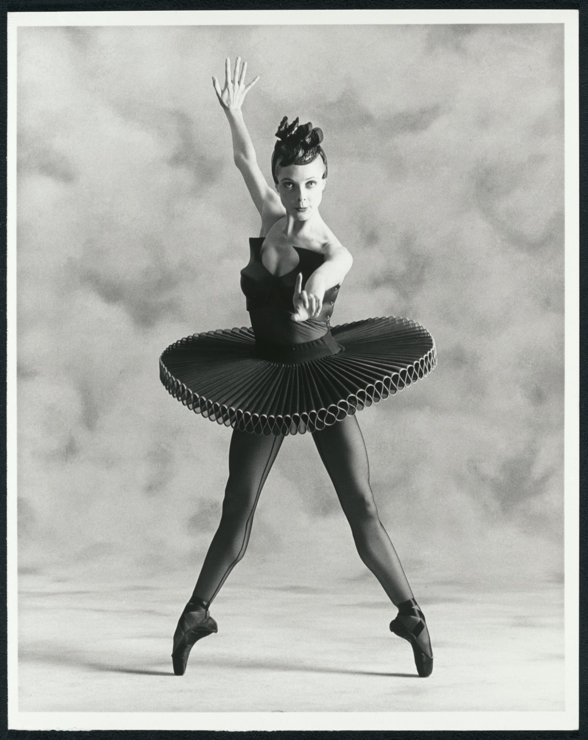 Ballerina Justine Summers strikes a pose for the camera. She is standing en point wearing a black tutu. She is standing infront of a textured curtain. She is staring directly at the camera.