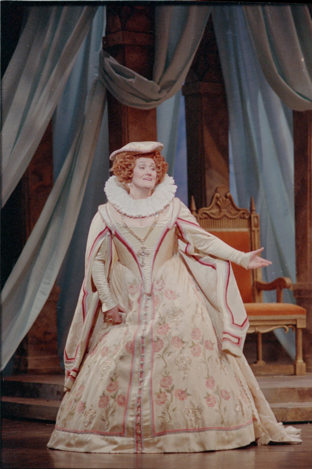 A picture of a woman on stage mid-performance. She is dressed in a large old-fashioned gown with ruffles. Behind her is a throne and heavy curtains