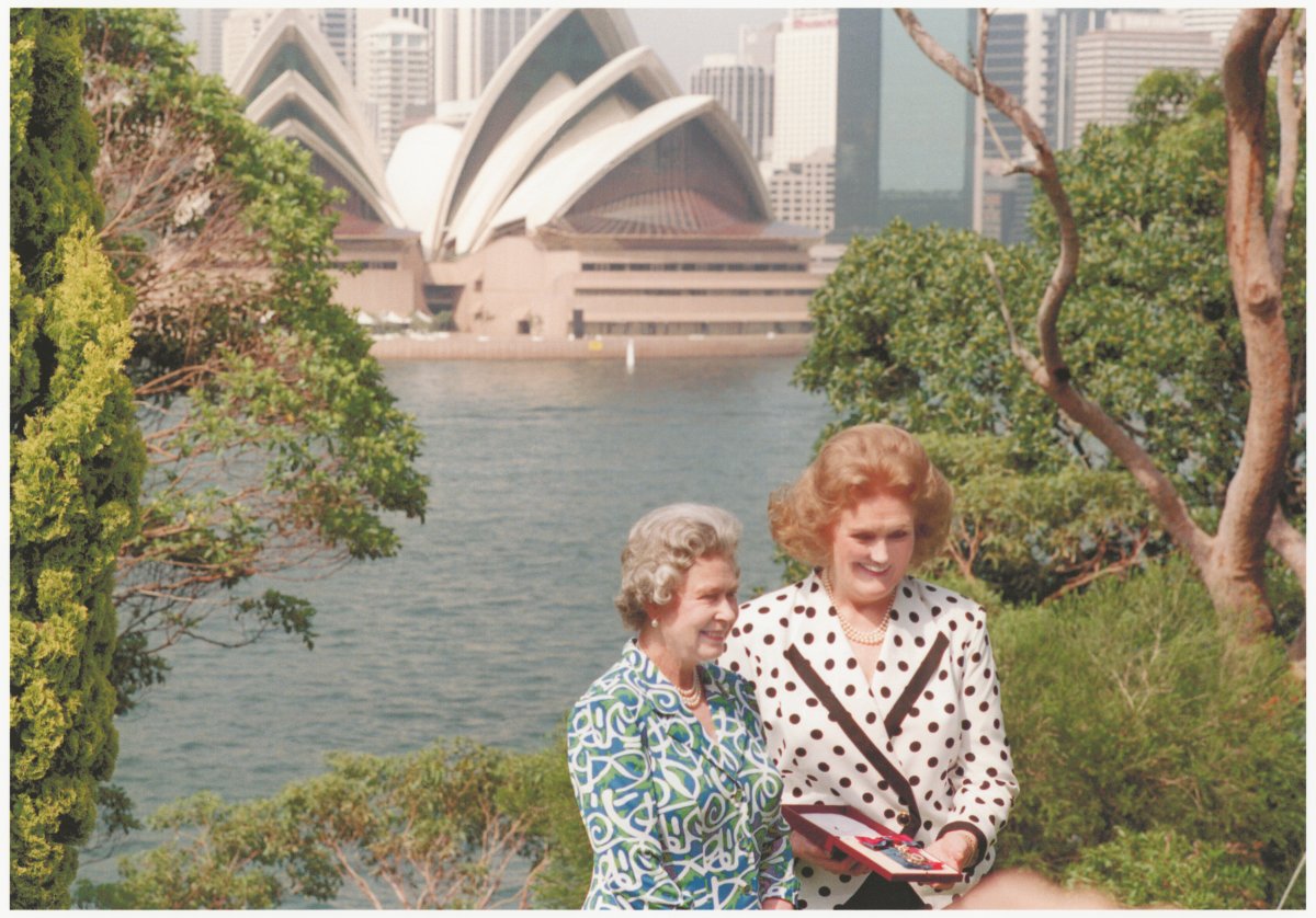 Two women stand among trees with the Sydney Opera House in the background. One lady wears a white and black polka dot blazer and has red hair. The other wears a green, white and blue patterned dress. She has grey hair. Both are smiling