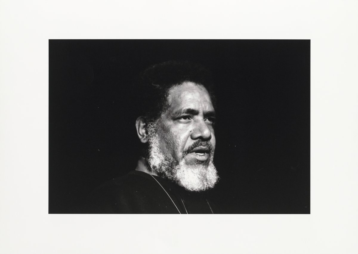 A black and white photograph of a man standing in front of a black background. He has a white beard and black hair. He is staring off camera mid-speech. The picture is surrounded by a thick white frame