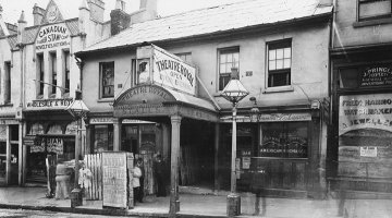 Black and white photograph of a row of shopfronts. One facade reads 'Theatre Royal'.