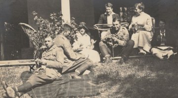 A sepia image of 7 people sitting on a porch and on the grass, knitting and winding wool