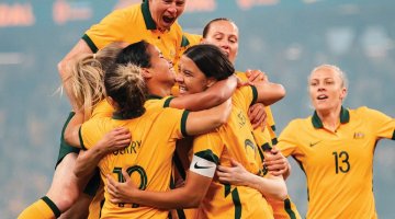 Seven women in the Australian soccer uniform all cheering and hugging one another.