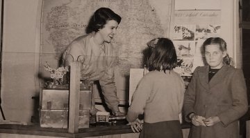 A sepia image of a lady behind a counter talking to two young girls who are standing in front of the counter.
