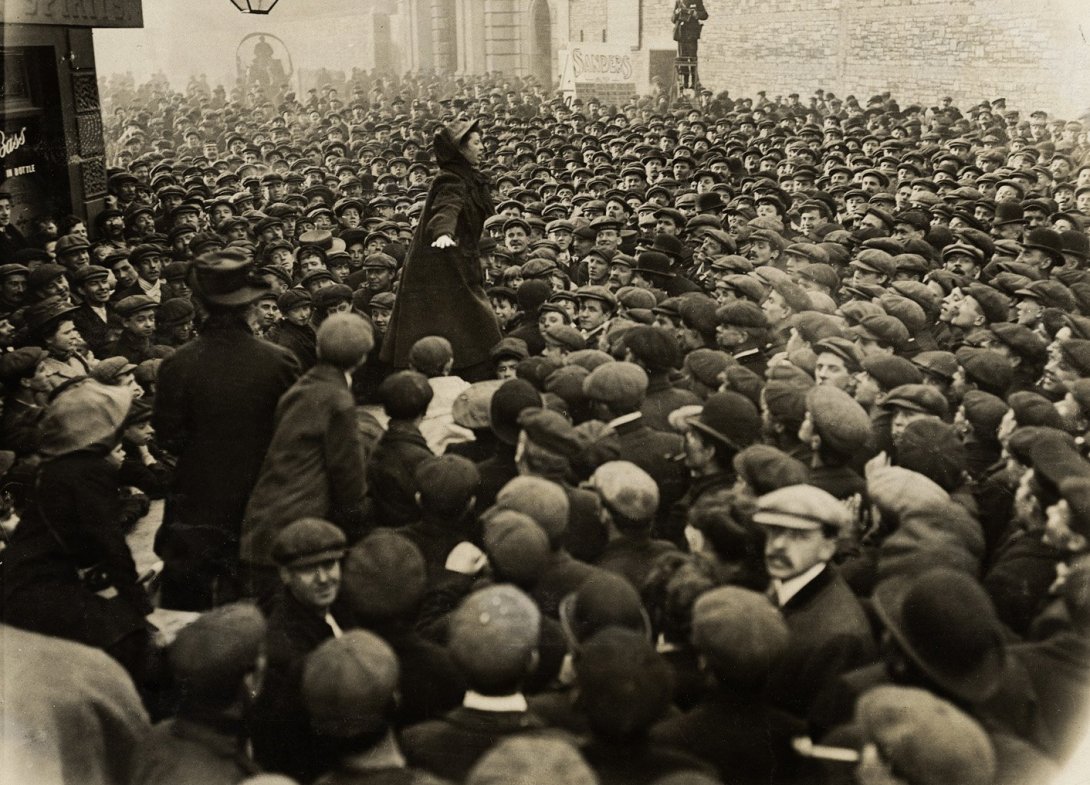 A woman standing with her arms stretched out speaking to a crowd of men outside a factory