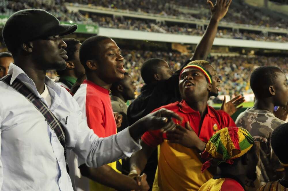 Three soccer fans, Ghana supporters, cheer on. Behind them can be seen a stadium full of spectators