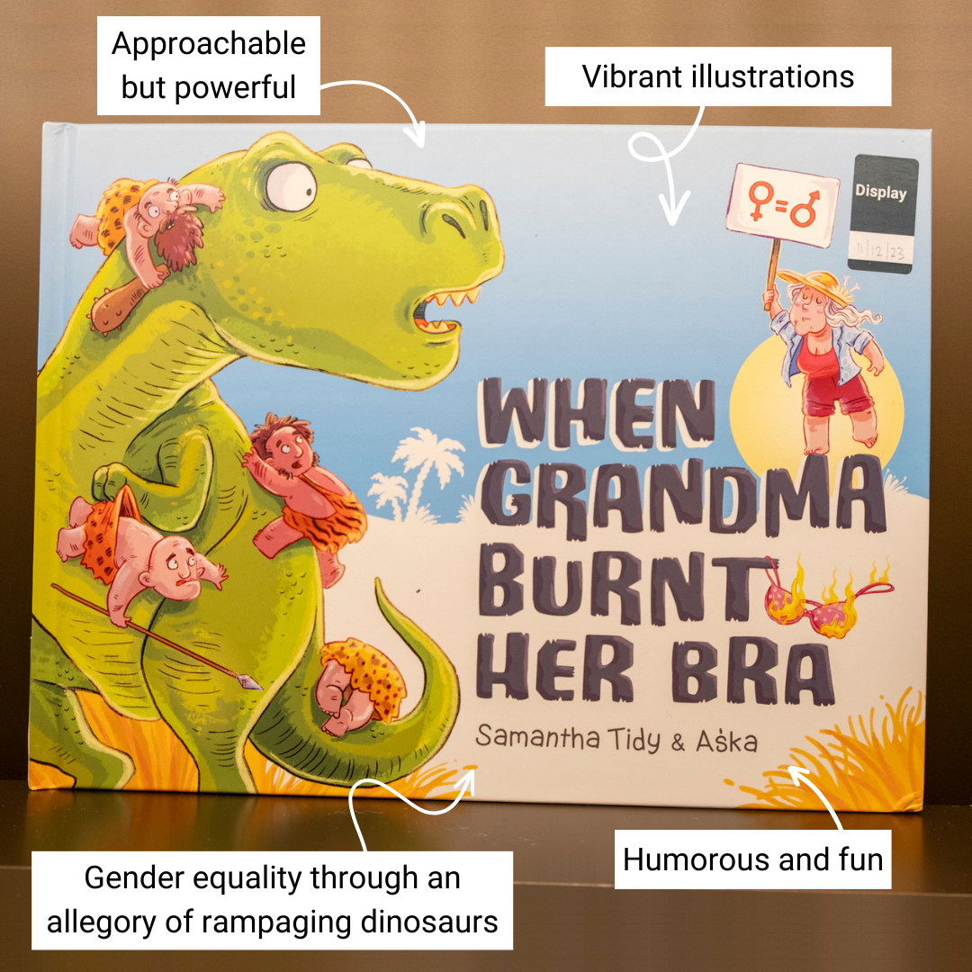 Children's book with an illustration of a dinosaur, cavemen and a grandma and text reading 'When Grandma Burnt Her Bra' and 'Samantha Tidy & Aska' on the cover sitting on display on a shelf. Annotations around the book read 'approachable but powerful', 'vibrant illustrations', 'gender equality through an allegory of rampaging dinosaurs' and 'humorous and fun'.
