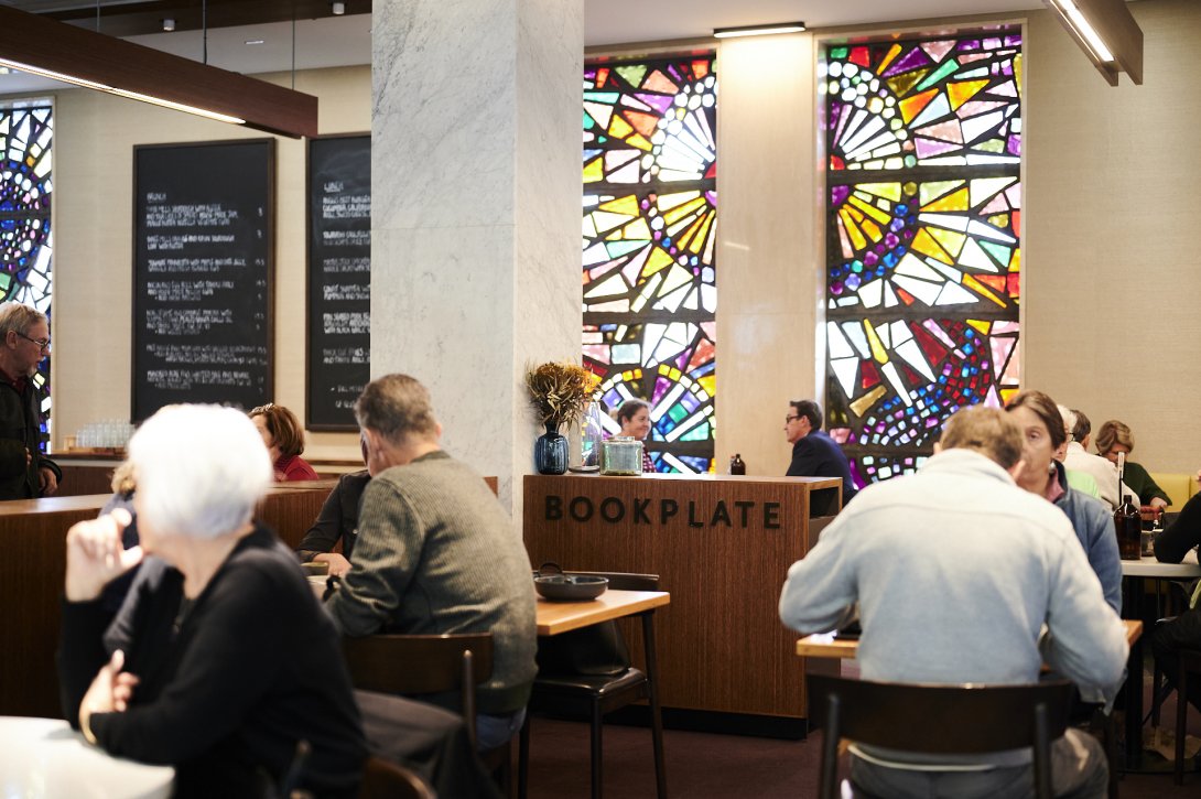 Cafe with people at every table enjoying food and coffee. On the far wall are large, colourful stained glass windows and large chalkboards with the menu. In the middle of the room is a wooden counter with 'BOOKPLATE' on the side