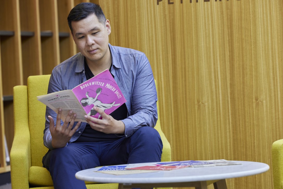 Man in blue button-up shirt and black hair sitting on a yellow armchair reading a booklet with a pink cover and text reading 'Bodenwieser Modern Ballet'