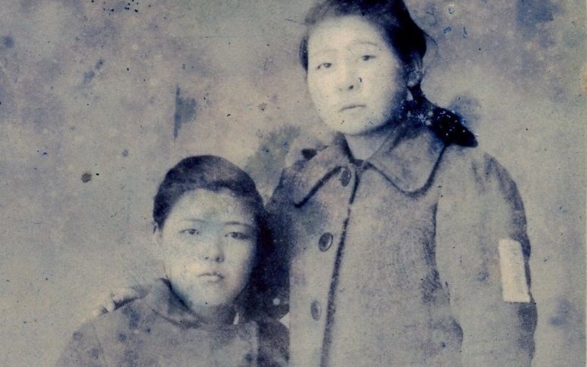 A black and white image of two women. One is seated and the other is standing with her arm around the seated woman.