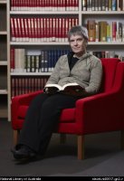 Sandra Wilson sitting in red chair in Asian Reading Room, bookcases behind her, a book open in her lap