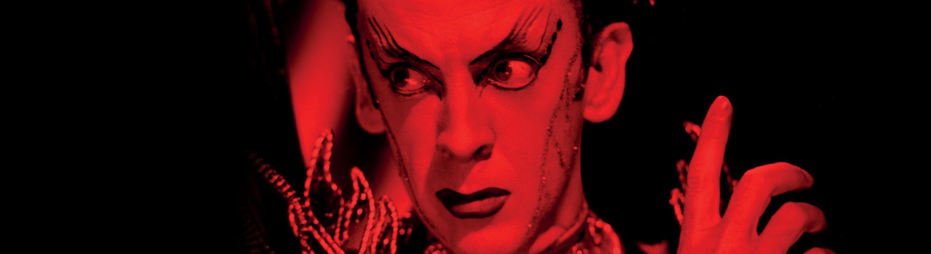 Robert Helpmann performs on stage with dramatic makeup. Their face is bathed in red light.