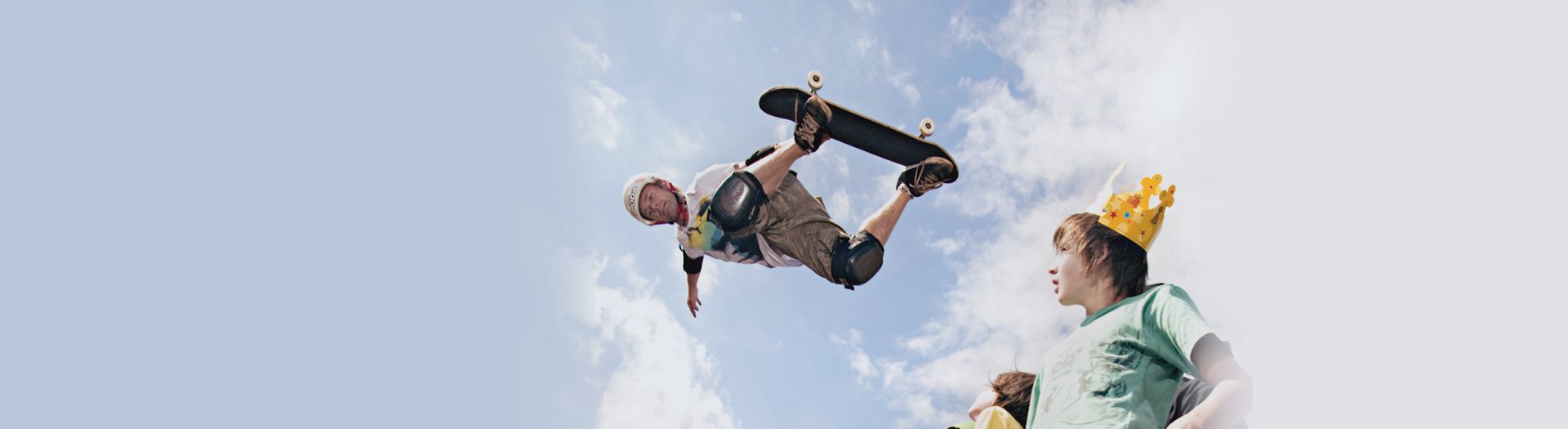The background is a blue sky with white clouds. A skateboarder is up in the air on his board. There are two people in the bottom right of the frame looking up at him.