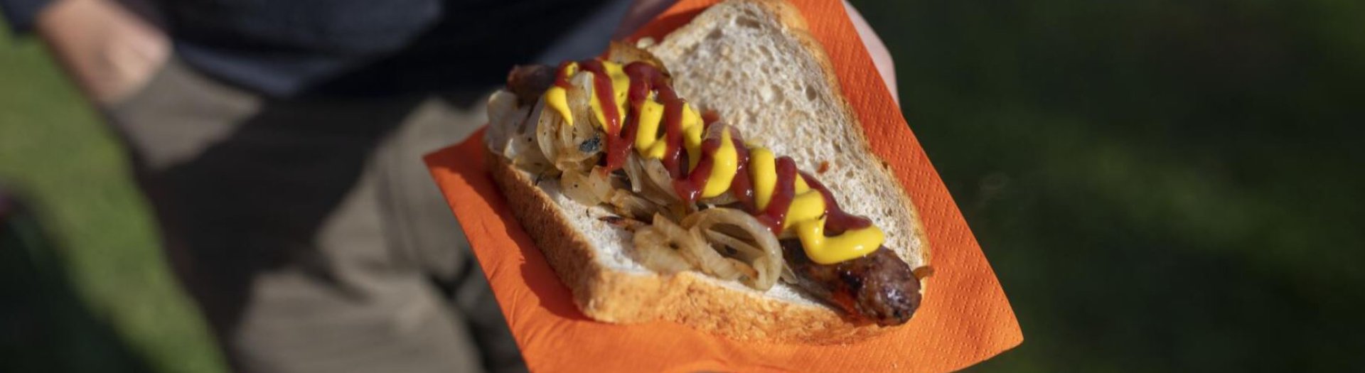 A sausage on a piece of white bread, covered in mustard, tomato sauce and fried onions. This sausage in bread is on an orange paper napkin, being held in someone's hand.