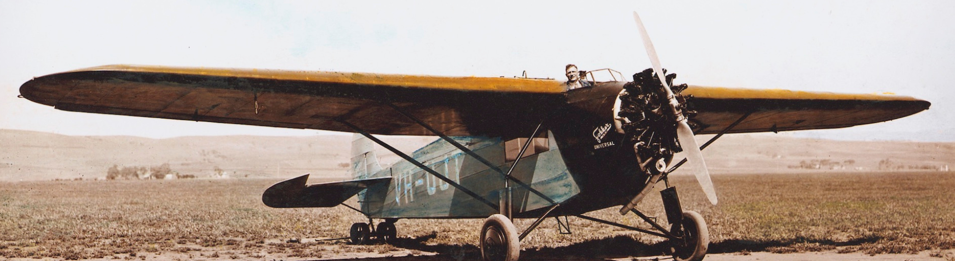 An old plane on the ground in a desert. A man is poking his head out from the cockpit and can be seen behind the wings of the plane