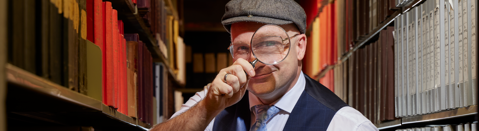 Man standing in the stacks of the Library wearing a button up shirt, tie, waistcoat, and newsboy cap holding a magnifying glass up to his eye with his right hand. 