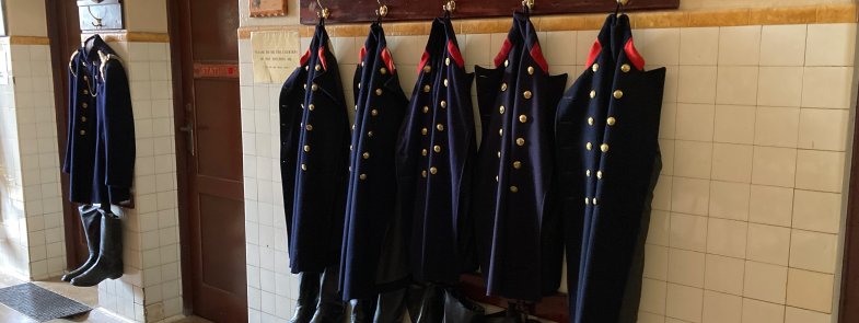 A row of firefighter uniforms with hats and boots are hung in a row on display