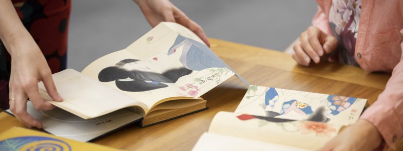 Two books open on a table with people flicking through them and looking at the books' colourful artwork showing drawings of women