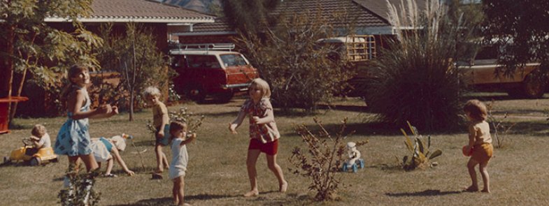 7 children playing in the front yard of a suburban house, throwing a colourful ball and driving toy cars