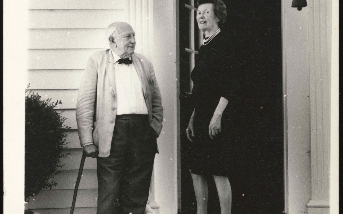 A black and white photo of an old balding man in a black bowtie, leaning on his cane and a woman in a black dress, smiling, about to enter a large white portal doorway, with a large black ship's bell hanging next to it.