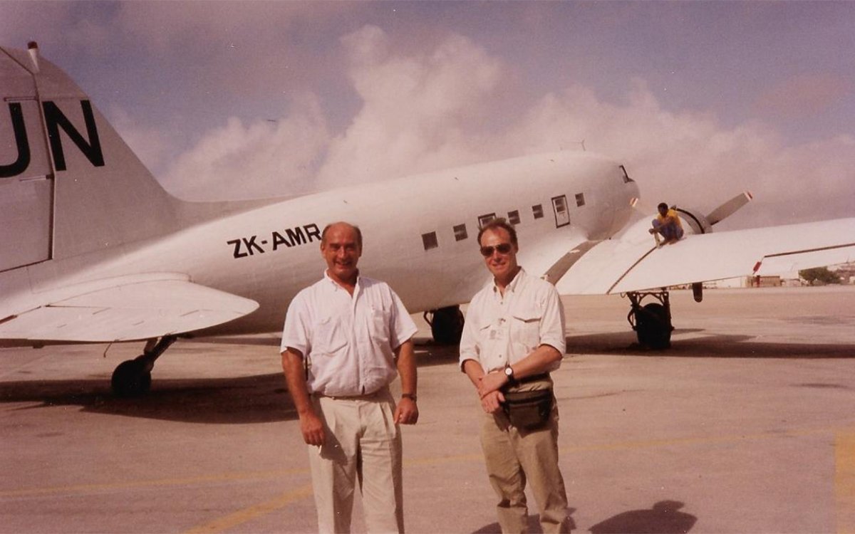Two men in white shirts and khaki pants stand in front of a small airplane