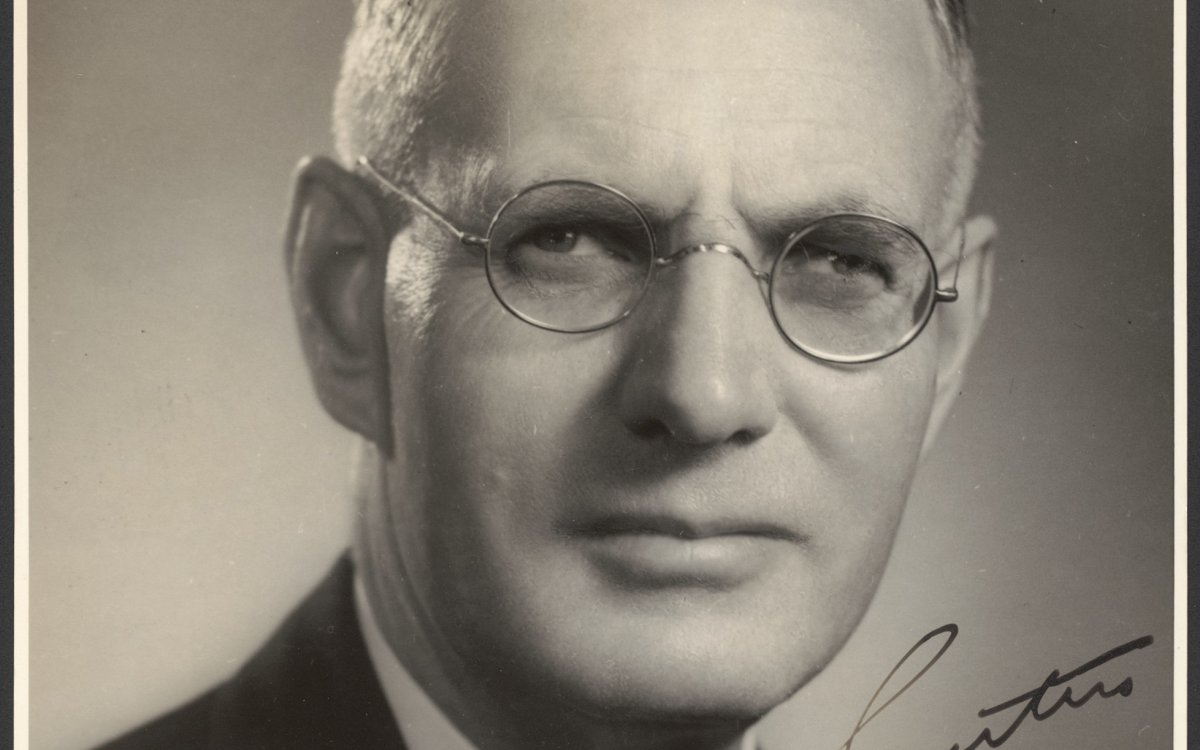 An official portrait of Australian Prime Minister John Curtain. He is a middle-aged white male wearing wire rimmed glasses. He has thinning blonde hair swept back neatly. He is wearing a dark suit and tie with a white collared shirt. He is staring directly at the camera with a stern look. In the lower right quadrant of the photograph is Curtains signature.