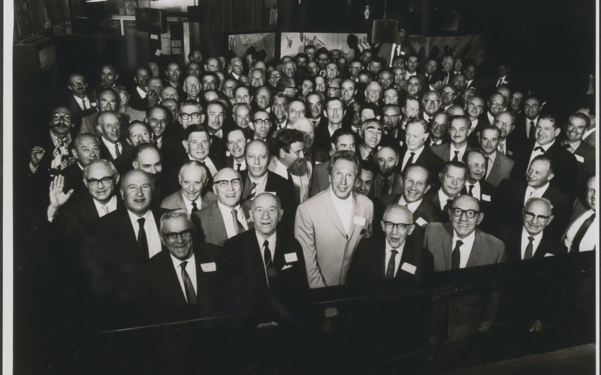 A black and white photograph of a large group of men, mainly older, standing in a large room. All the men are wearing dark suits and ties except one man in the centre of the photo. He is wearing a white suite with a white turtleneck sweater. Many of the men are wearing glasses. Some are waving at the camera and almost all are smiling broadly.