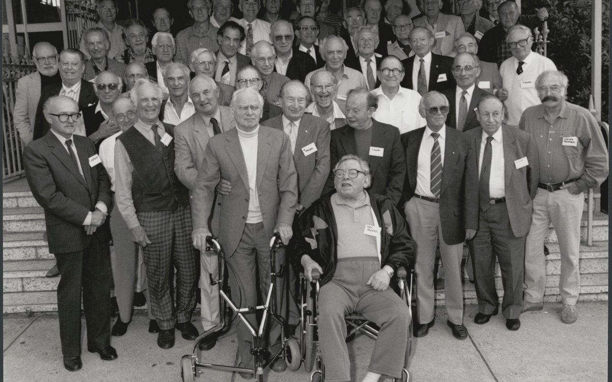 A black and white photograph of a large group of older men, posed for a group photo.  Most of the men are smiling at the camera.In the middle of the front row is a man sitting in a wheelchair. To his left a man stands holding a walking aide while a man holds his arm. 