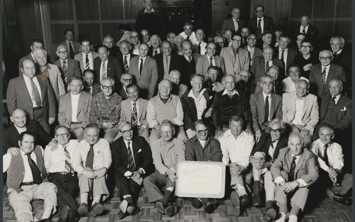 A black and white photograph of a large group of men, mainly older, posed for a group photo. Some men are sitting in the front rows, others stand at the back. They are all smiling at the camera. A man in the centre of the from row hold a white sign with black writing. It reads "Dunera Reunion 1981"