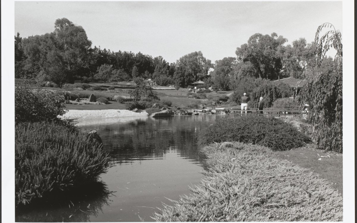 A black and white photograph of a manicured Japanese garden. There is a large calm lake in the centre of the image. There are large trees in the background and shorter shrubbery in the foreground. Two people are standing on the shore of the lake looking away from the camera.