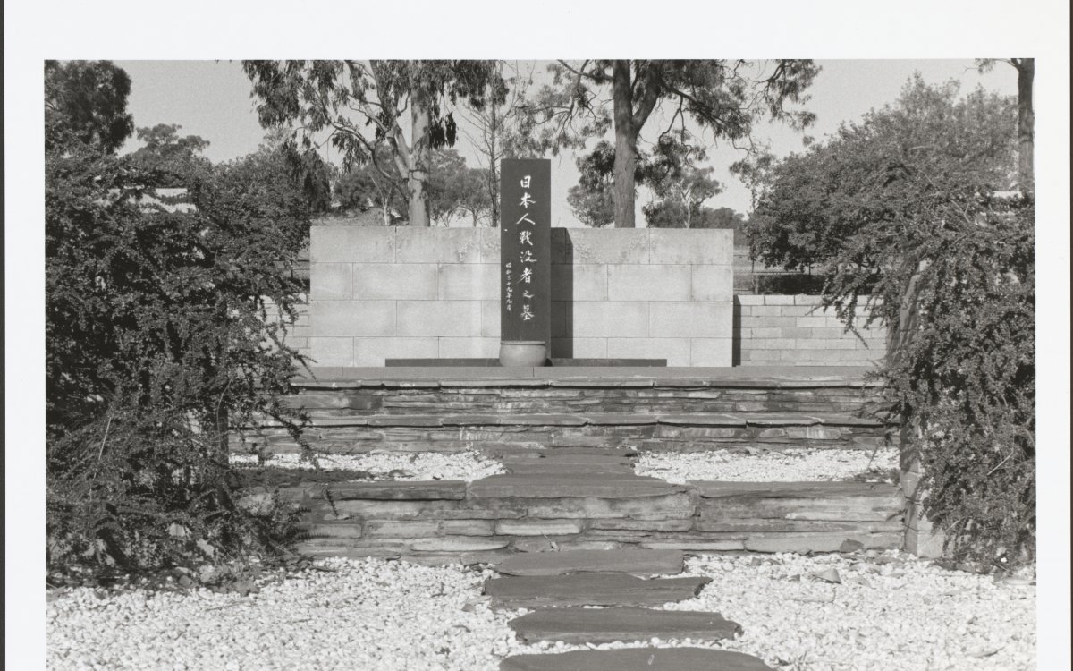 A black and white photograph of a stone memorial. In the middle of the memorial is a black stone obelisk with carved Japanese kanji. The site is surrounded by trees. There is a flagstone path surrounded by white pebbles leading up to the memorial