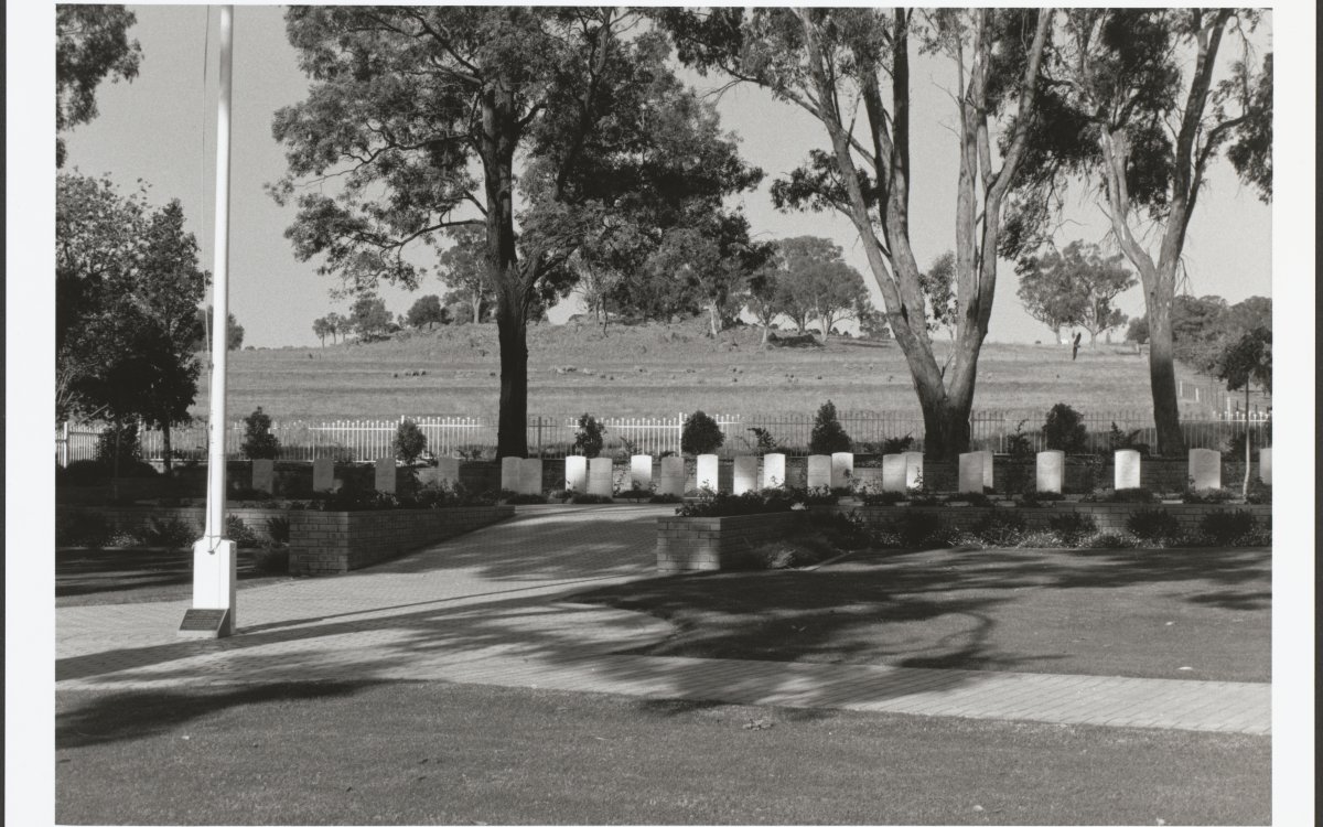 A black and white photograph of a memorial area. There are a row of 23 white headstones along the middle of the image. They are in the shade of large gumtrees. There is a white flagpole on the left of the image. In the background there is a grassy hill.
