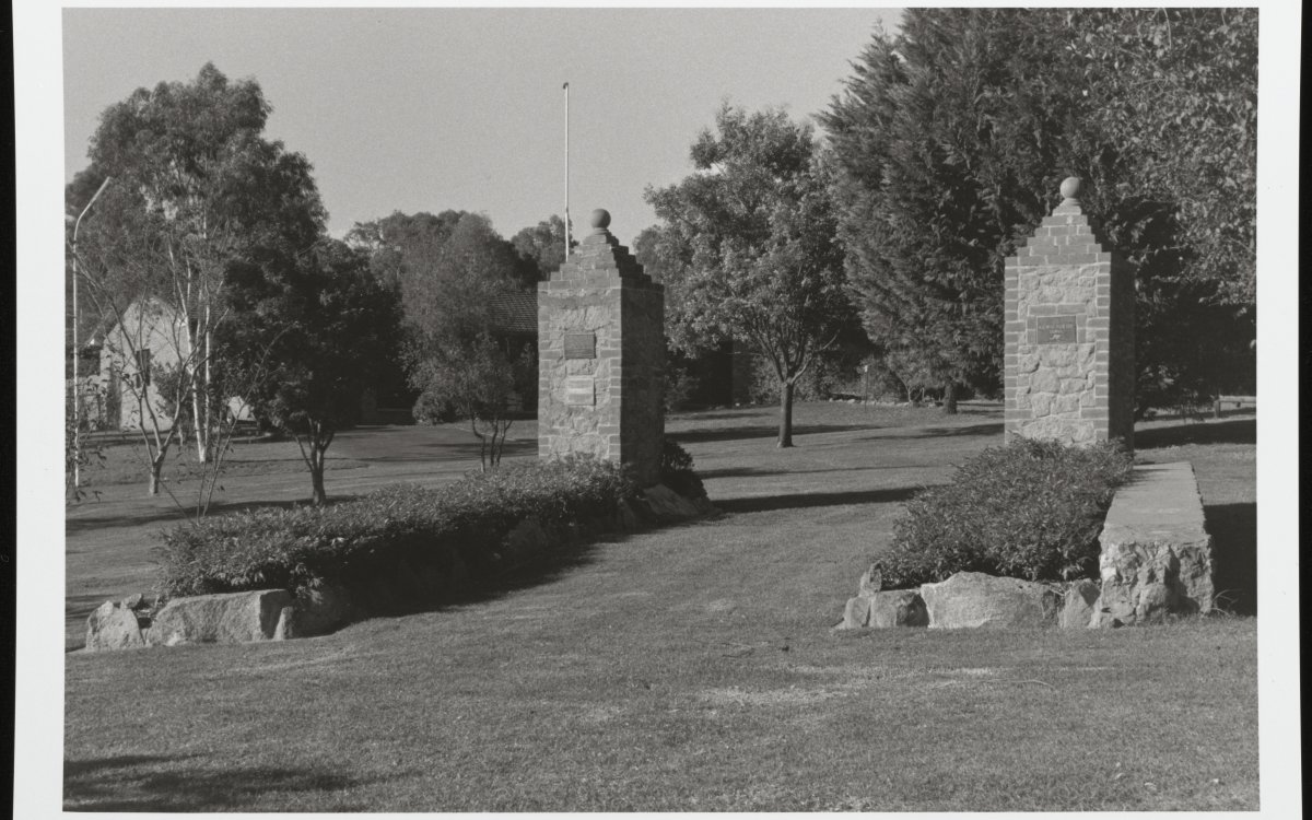 A black and white photograph of the main garrison gates of the former Cowra Prisoner of War camp. The gates now sit in a grassy park surrounded by trees and bushes.