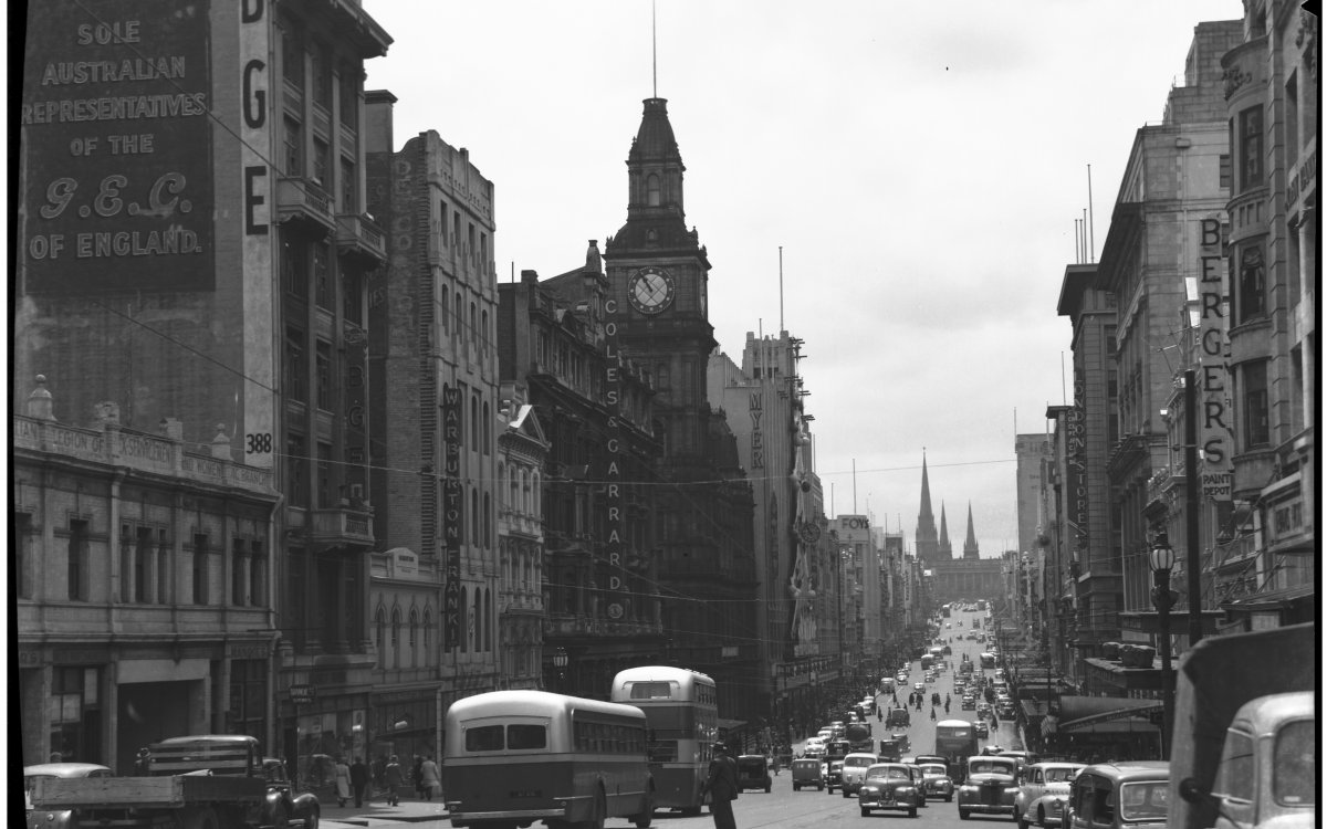 	 Bourke Street looking east; day time view photographed in the late 1940s or early 1950s. Two buses are prominent int he foreground, followed by a flatbed lorry. A large clock tower dominates the center of the image. A church steeple can be seen int he background.