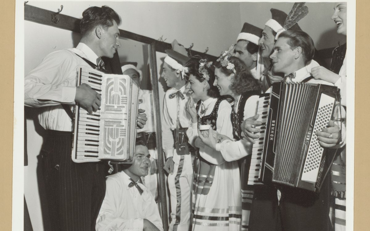 A group of people stand around two men playing button accordians. Some people are wearing Polish traditional dress others are in shirts and black pants. They look to be mid-song. Everyone is smiling.