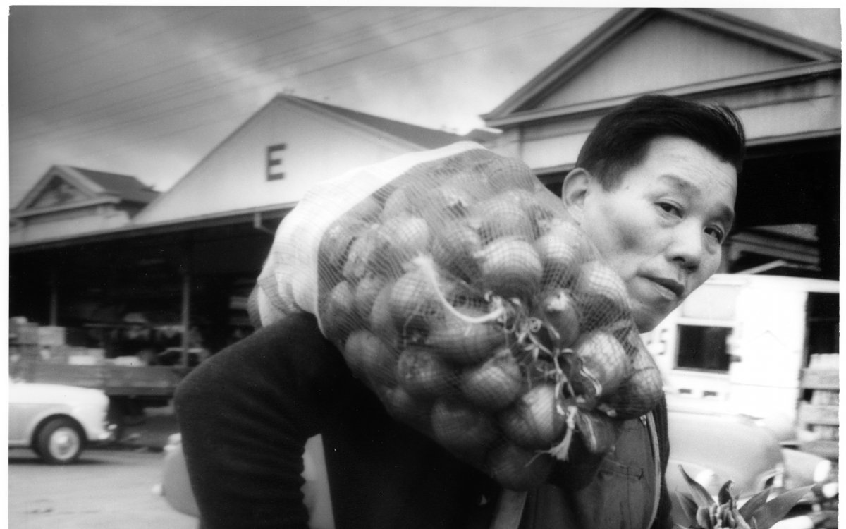 A man carriees a bag of onions over his shoulder. Behind him are the sheds of the Queen Victoria Markets, a large produce market. The man is wearing dark overalls and two cardigans