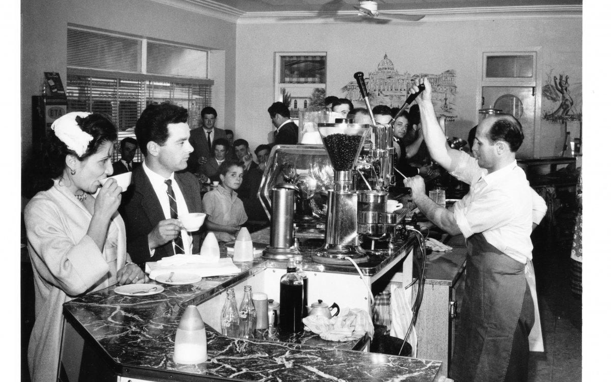 Image of group of people eating and drinking in a coffee shop. A man behind the counter operates a gleaming coffee machine.