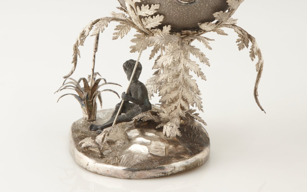 A sculpture of an emu egg on Silver-plate Stand. Underneath an Aboriginal man sits with spear. Surrounding the figure are large fern fronds and rocks