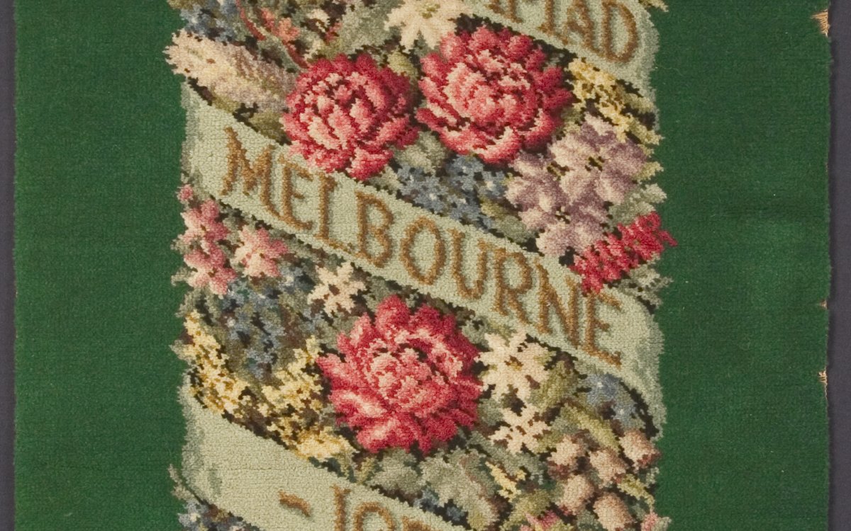 A green carpet square with a floral motif and writing embroidered on it. The woven design features an Australian motif of red waratahs, blue leschenaultias, flowering gums, white flannel flowers and the bottlebrush The writing says "XVI (16) Olympiad Melbourne 1956". 