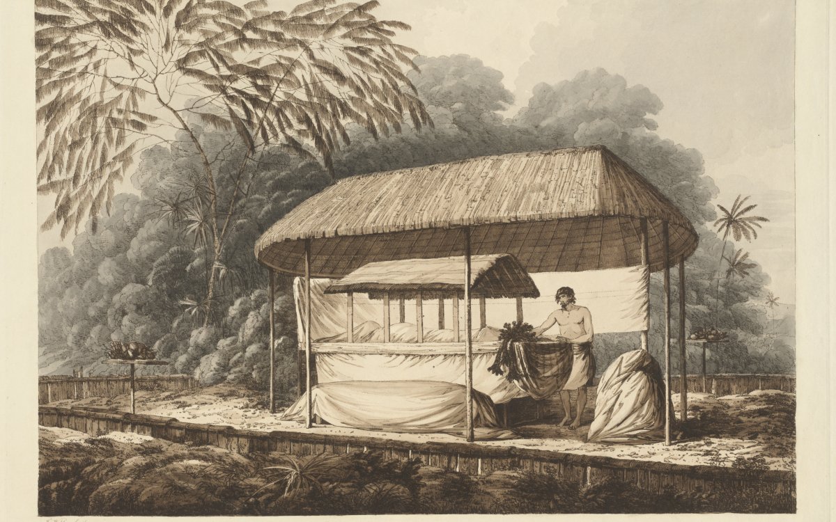 A man stands next to a ceremonial alter. The alter has a thatched roof. It sits inside a larger shelter also with a thatched room. On the alter is a body shrouded in cloth. Behind the shelter is a large thicket of trees.