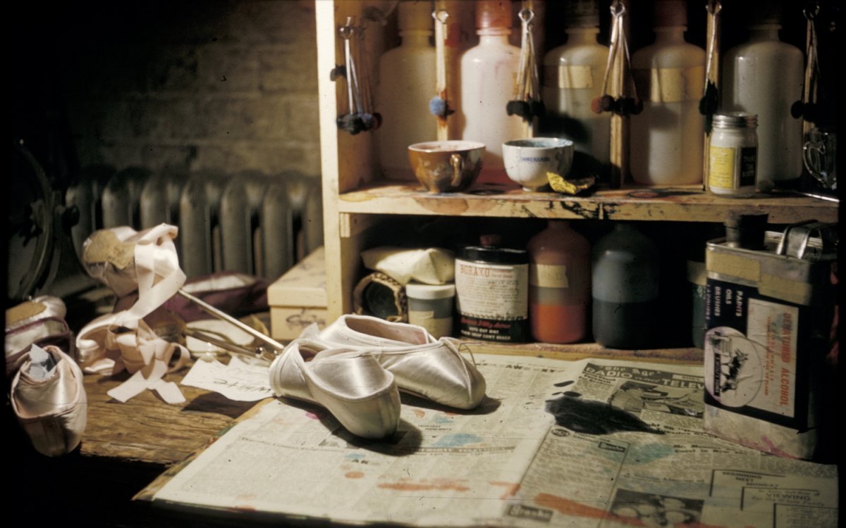 Ballet slippers are sitting on newspaper on a table with paints and other craft items on a shelf behind.