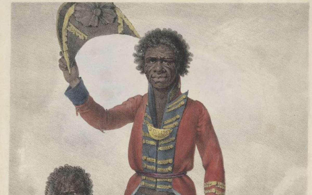 A portrait of a man. He has dark skin and hair. He is an Australian Indigenous man and is wearing a red, blue and gold European style officers coat and trousers. His trousers are tattered at the cuffs. He is holding a tricorn hat in the air. In the background an Indigenous women is seated, wrapped in a shawl. She is smoking a pipe. There is a woven basket with two empty bottles. In the background there is a white washed house with smoke curling from the chimney.