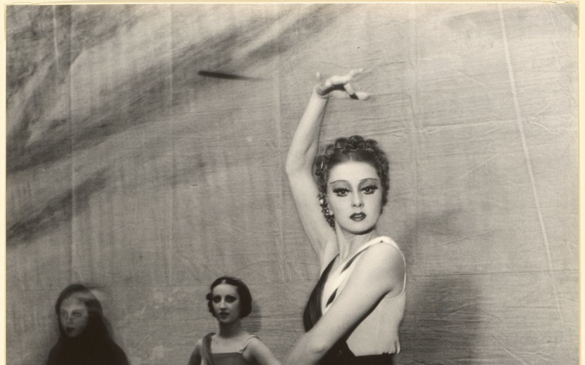 Ballerina Irina Baronova strikes a ballet pose for the camera. She is standing en point wearing a long black dress. Two dancers are watching her in the background