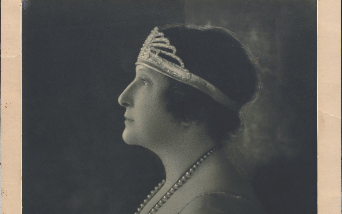 A formal portrait photograph of a lady wearing a diamond tiara. She is in profile. She has short hair and a lace gown on. She is wearing a long string of pearls. The photograph is signed "Nellie Melba 1922"