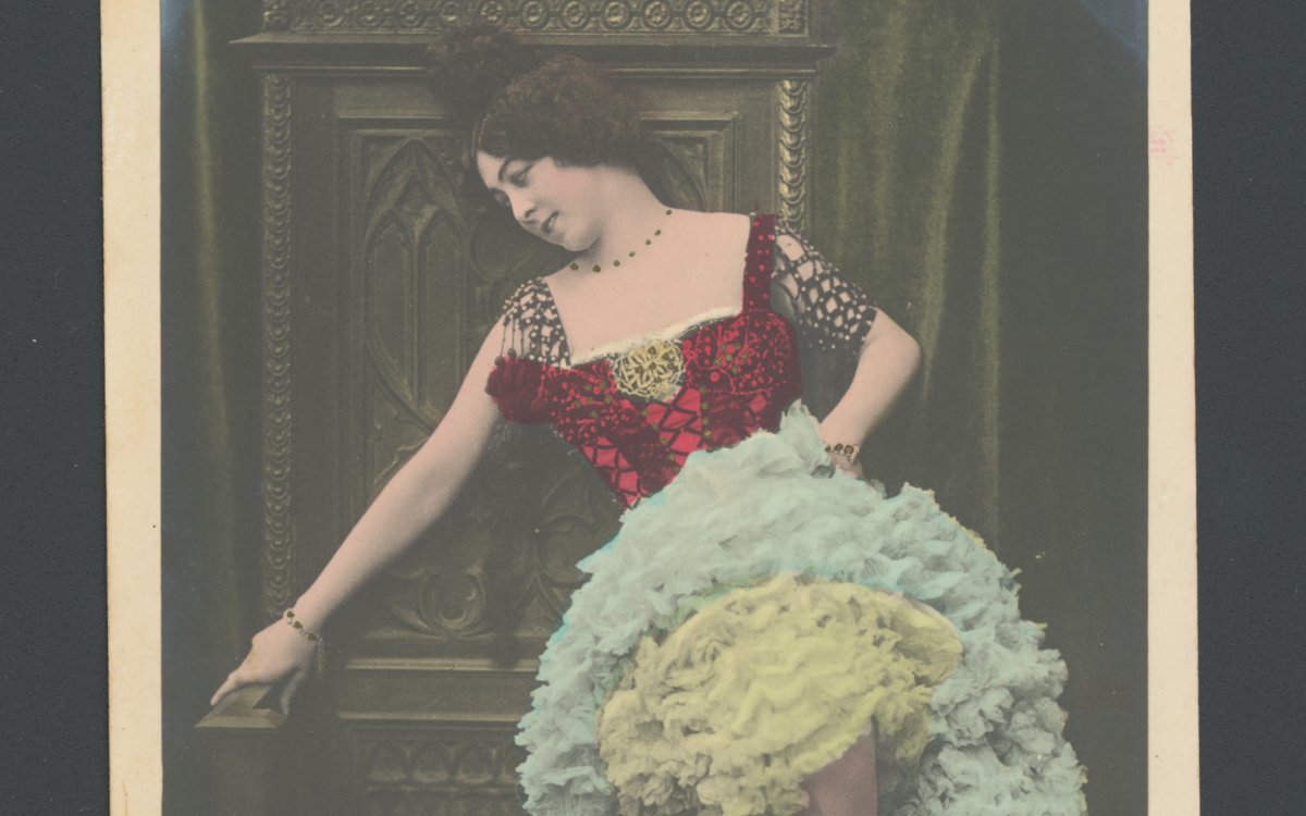 A vintage colourised photograph of a woman sitting on a the arm of a large throne or chair. She is wearing a dress with a red bodice and multiple layers of frills in blues and yellows. She has ballet shoes on. On the floor in a wreath.
